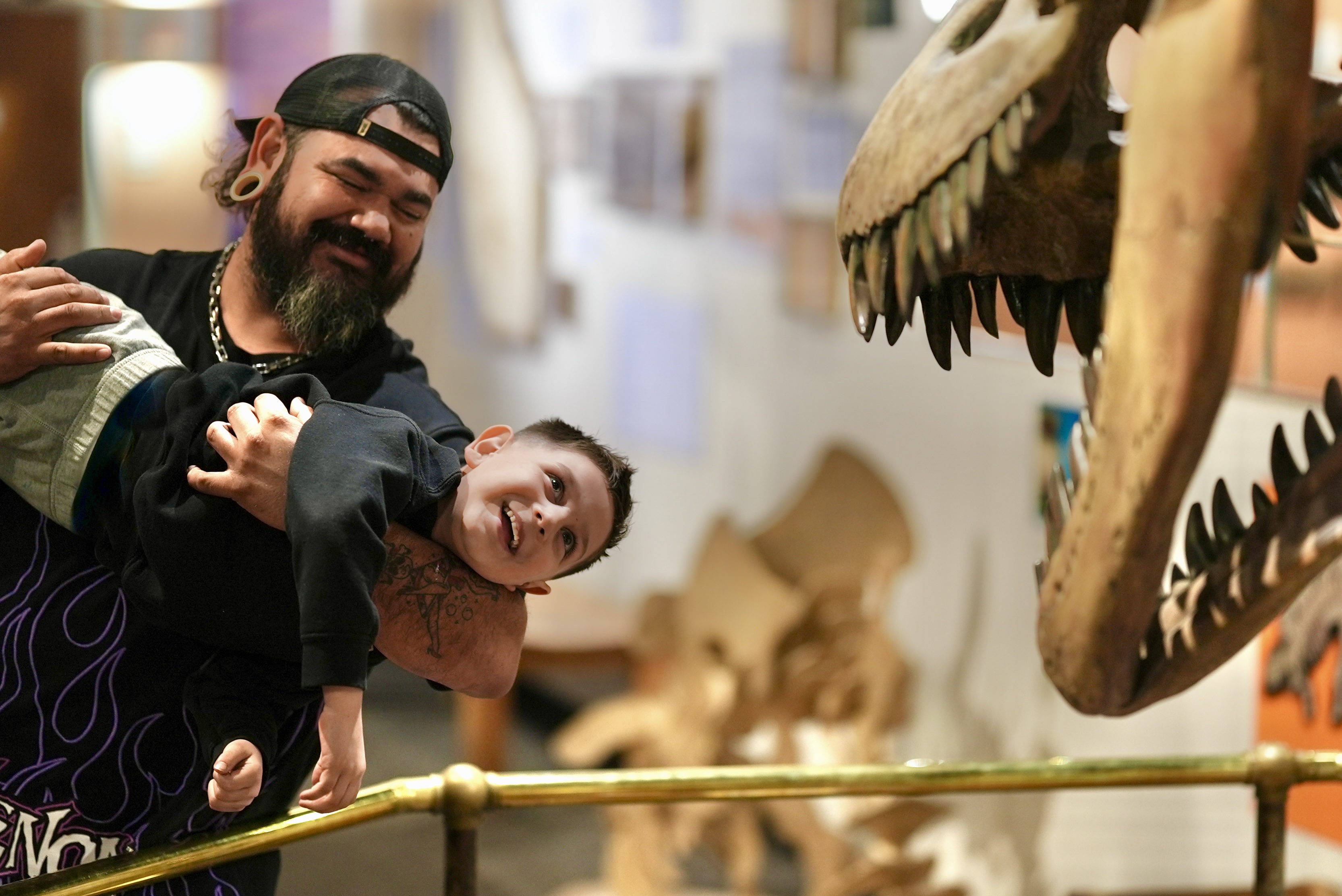 A man holds a child, they both have big smiles as the man pretends to feed the child to the dinosaur!